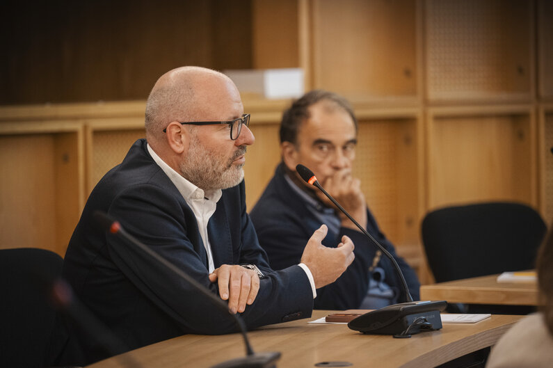 Ein Mann sitzt an einem Tisch. Er spricht in ein Mikrofon.