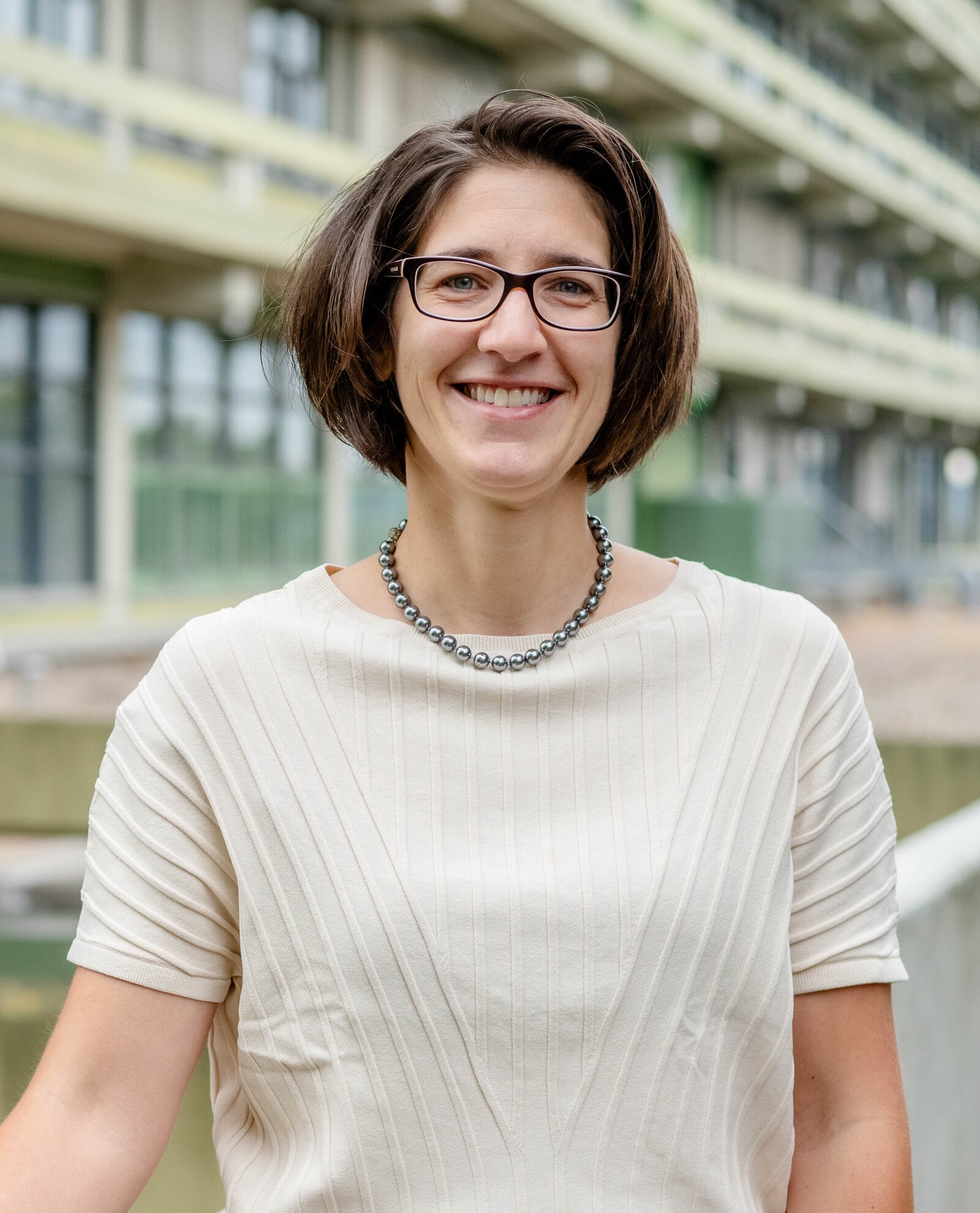 Eine Frau mit Brille und halblangen Haaren steht lächelnd vor einem Gebäude.