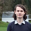 Ein Mann mit langen dunklen Haaren steht in einem Garten und sieht lächelnd in die Kamera. 