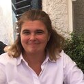 Eine Frau steht vor einem Haus, hinter ihr ist ein Fenster und ein Schlagladen aus Holz zu sehen. Sie lächelt in die Kamera.