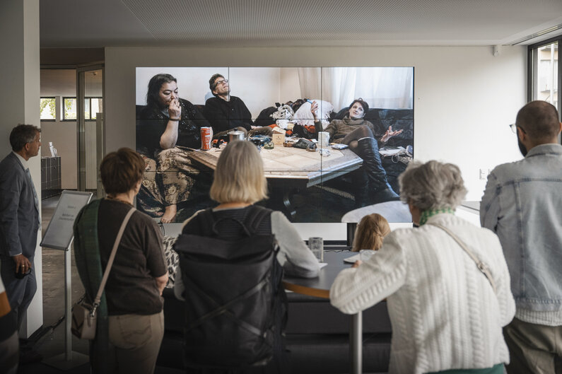 Eine Gruppe von Menschen schaut auf eine Leinwand. Auf dieser sind drei Personen, die auf einem Sofa sitzen, zu sehen.
