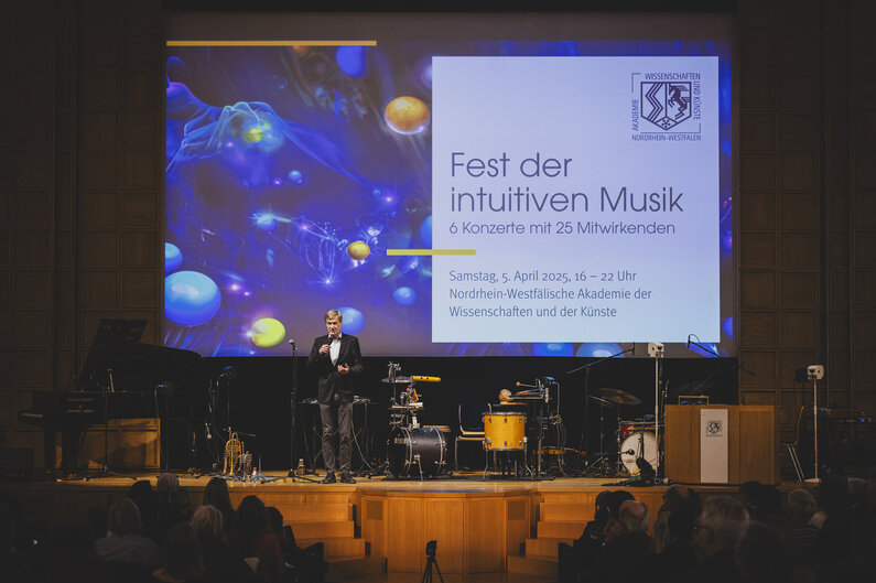 Ein Mann steht auf einer Bühne. Auf einem Bildschirm ist der Titel der Veranstaltung eingeblendet: Fest der intuitiven Musik.