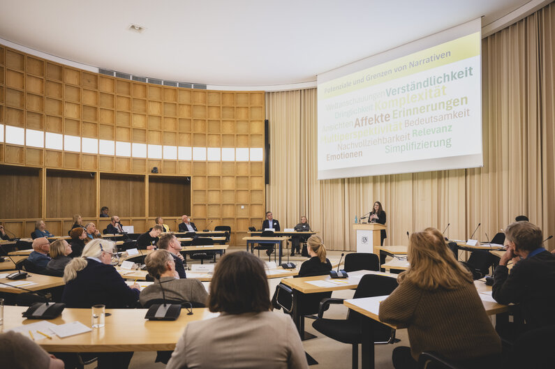 Männer und Frauen sitzen in einem halbrunden Saal an Tischen. Vorne steht eine Frau an einem Rednerpult.