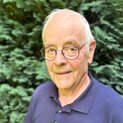 Ein Mann mit Brille steht lächelnd vor einem Strauch.